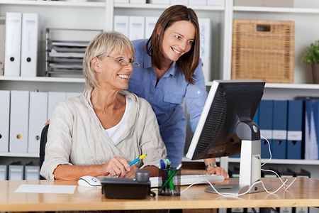 Zwei Frauen im B&uuml;ro - eine &Auml;ltere sitzt vor dem PC, die J&uuml;ngere steht hinter ihr und schaut ihr &uuml;ber die Schulter.