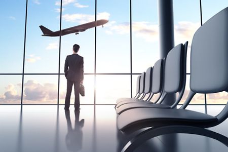 Mann steht in Anzug und mit Aktentasche am Fenster des Flughafengeb&auml;udes und schaut einem startenden Flugzeug zu