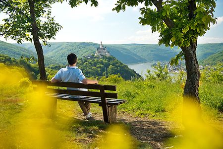 Wanderer sitzt auf Bank mit Blick auf Rhein und Marksburg
